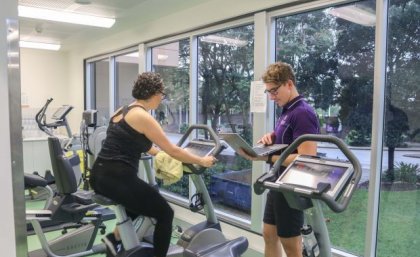 Supervised exercise sessions will be delivered via a videoconferencing platform. Supplied. Woman on exercise bike while man stands nearby with laptop. Supplied.